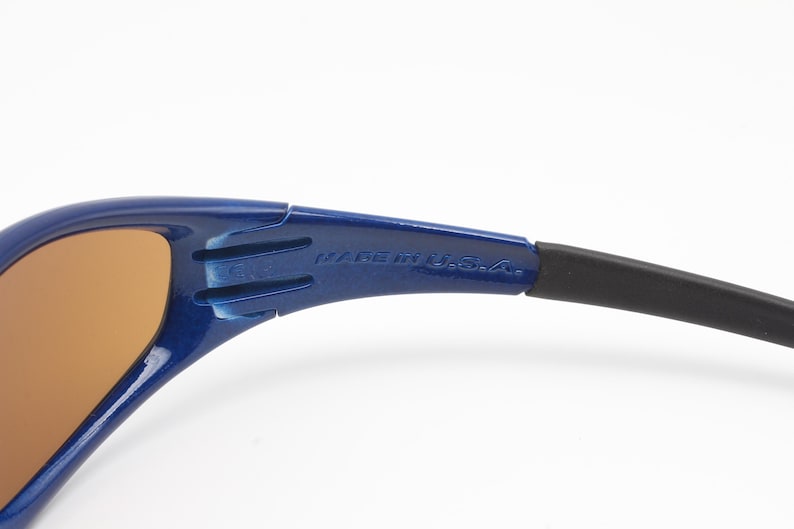 May include: Close-up of a blue and black pair of sunglasses with brown lenses. The sunglasses have the text "Made in U.S.A." printed on the side.