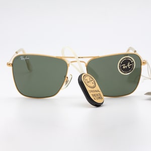 May include: Gold-framed sunglasses with green lenses. The sunglasses have a Ray-Ban logo on the side and a "Original Bausch & Lomb" sticker on the arm.