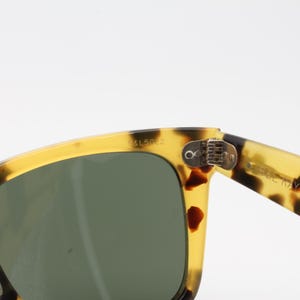 Puede incluir: Primer plano de gafas de sol con montura con estampado de carey y lentes verde oscuro. La montura es una mezcla de amarillo y marr&oacute;n, con el texto "B15022" visible en la esquina superior izquierda. La patilla de las gafas tiene el texto "MADE IN ITALY" y "GAN S.A."