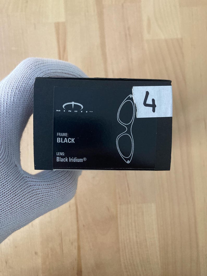 May include: Black eyewear box with the text "MINUTE" and a diagram of the glasses. The box also has the text "FRAME: BLACK" and "LENS: Black Iridium&reg;". A white sticker with the number "4" is on the box.