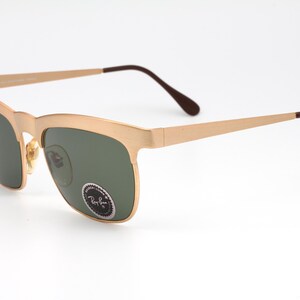 May include: Gold-tone Ray-Ban sunglasses with green lenses. The sunglasses have a square frame with a slightly rounded edge.