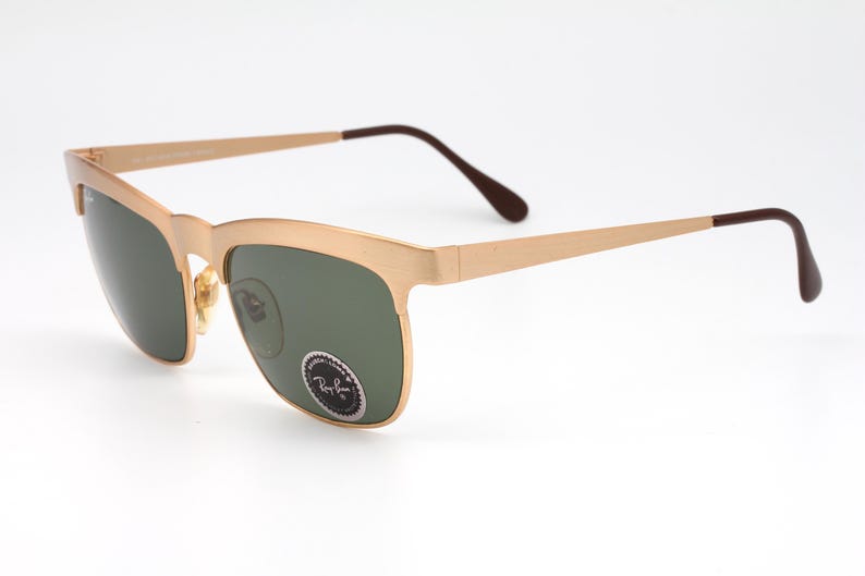 May include: Gold-tone Ray-Ban sunglasses with green lenses. The sunglasses have a square frame with a slightly rounded edge.