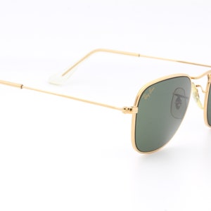 May include: Gold-framed square sunglasses with green lenses. The Ray-Ban logo is visible on the right lens.