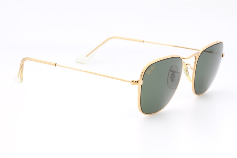 May include: Gold-framed square sunglasses with green lenses. The Ray-Ban logo is visible on the right lens.