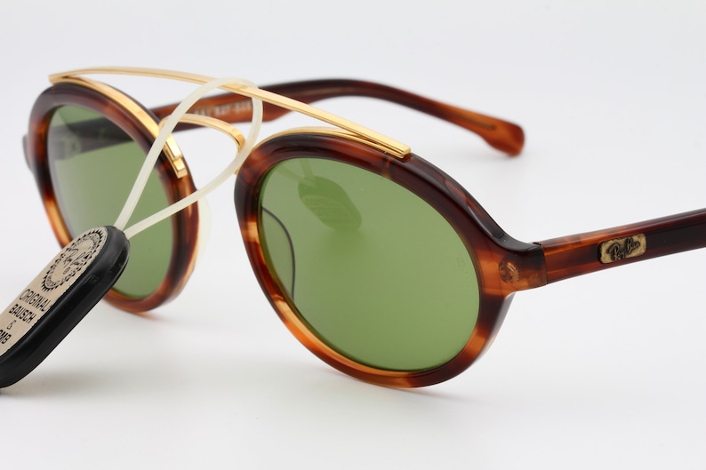 May include: Tortoise shell frame sunglasses with green lenses and a gold metal bridge. The sunglasses have a Ray-Ban logo on the side.
