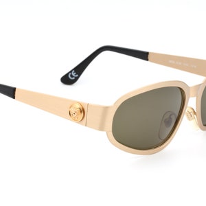 May include: Gold-toned sunglasses with brown lenses and a black Versace logo on the side. The sunglasses have a black temple and a gold-toned frame.
