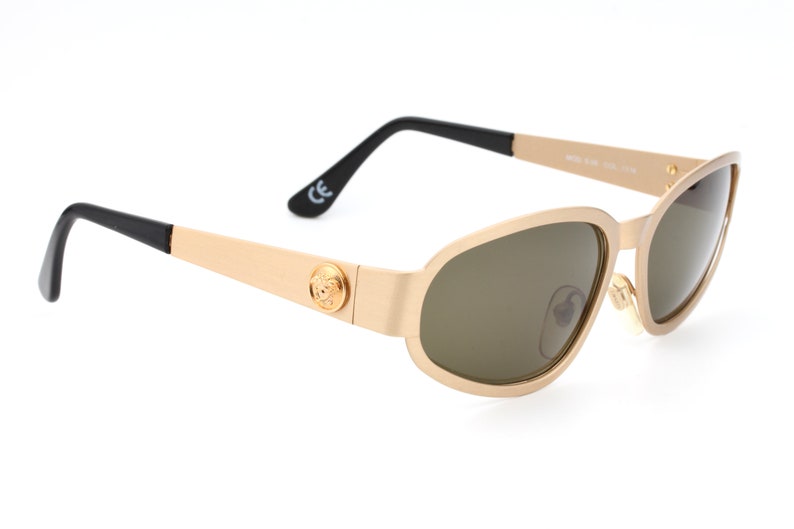 May include: Gold-toned sunglasses with brown lenses and a black Versace logo on the side. The sunglasses have a black temple and a gold-toned frame.