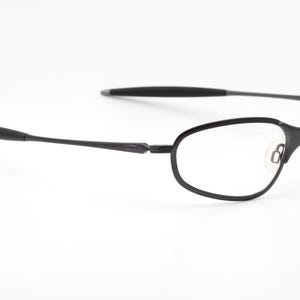May include: Black eyeglasses with a minimalist design. The frames are a matte black, with a thin, oval shape around the lenses. The temples are slender and extend to the ear pieces. The glasses are set against a white background.