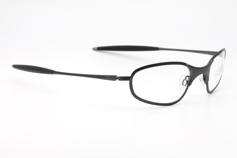 May include: Black eyeglasses with a minimalist design. The frames are a matte black, with a thin, oval shape around the lenses. The temples are slender and extend to the ear pieces. The glasses are set against a white background.
