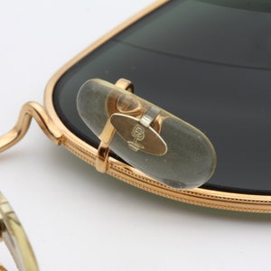 May include: Close-up of gold-tone metal sunglasses with a clear plastic nose bridge and dark green lenses. The sunglasses have a B&L logo on the nose bridge.