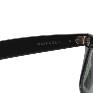 Puede incluir: Gafas de sol Wayfarer negras con bisagra plateada. Las gafas tienen lentes verde oscuro.
