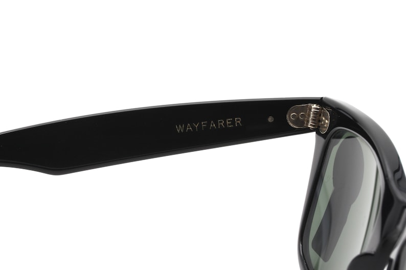 Puede incluir: Gafas de sol Wayfarer negras con bisagra plateada. Las gafas tienen lentes verde oscuro.