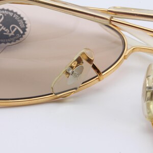 May include: Gold-tone aviator sunglasses with brown lenses. The sunglasses feature a Ray-Ban logo on the left lens and a "CHANGEABLE" label on the left arm. The sunglasses have a white plastic frame.