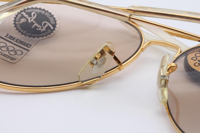 May include: Gold-tone aviator sunglasses with brown lenses. The sunglasses feature a Ray-Ban logo on the left lens and a "CHANGEABLE" label on the left arm. The sunglasses have a white plastic frame.