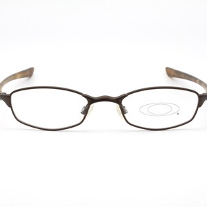 May include: Brown metal-framed eyeglasses with oval lenses. The temples have a patterned design. The Oakley logo is visible on the right lens. The glasses are set against a white background.