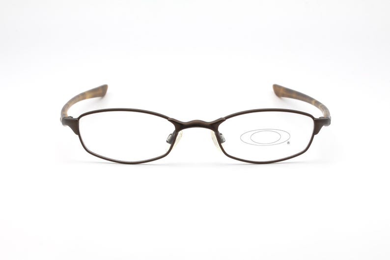 May include: Brown metal-framed eyeglasses with oval lenses. The temples have a patterned design. The Oakley logo is visible on the right lens. The glasses are set against a white background.