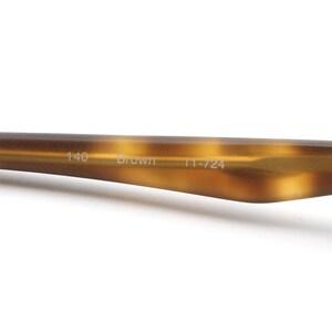 May include: Close-up of a brown eyeglass arm with a tortoiseshell pattern. The text "140 Brown 11-724" is printed on the arm. The arm is made of a glossy material and is set against a white background.