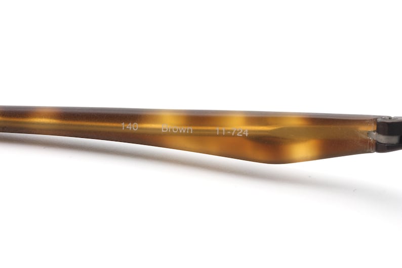 May include: Close-up of a brown eyeglass arm with a tortoiseshell pattern. The text "140 Brown 11-724" is printed on the arm. The arm is made of a glossy material and is set against a white background.