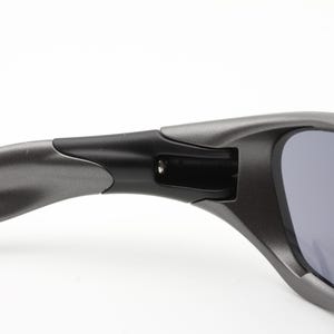Może przedstawiać: Zbliżenie na parę szarych okular&oacute;w przeciwsłonecznych. Okulary przeciwsłoneczne mają matowe wykończenie i elegancki, nowoczesny design. Soczewki są ciemnoszare, a oprawka jaśniejsza. Zawias jest czarny.