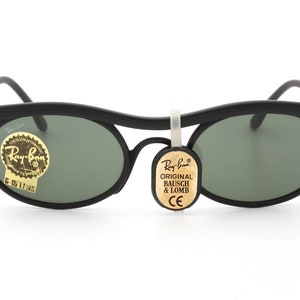 May include: Black oval-shaped sunglasses with green lenses. The sunglasses have a thin black frame and a Ray-Ban logo on the side. The sunglasses are attached to a tag that reads "Ray-Ban Original Bausch & Lomb CE".
