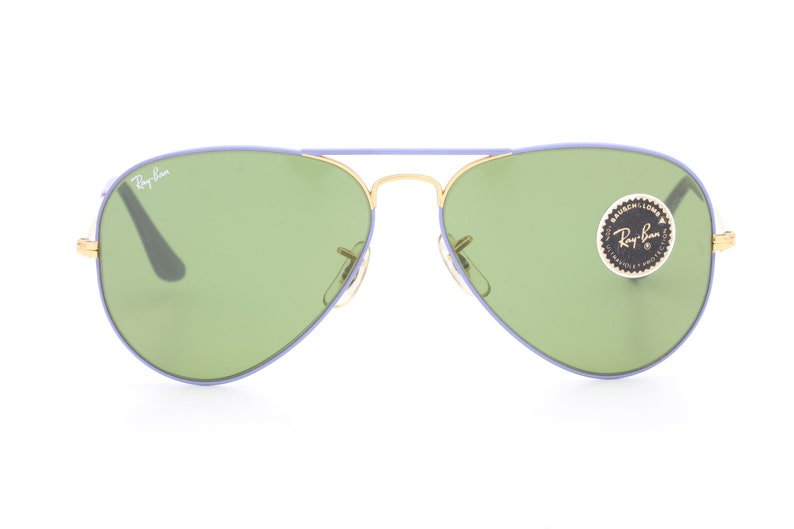 Op de afbeelding: Paarse en gouden aviator zonnebril met groene glazen. De zonnebril heeft een Ray-Ban logo op de linkerlens en een Ray-Ban sticker op de rechterlens.