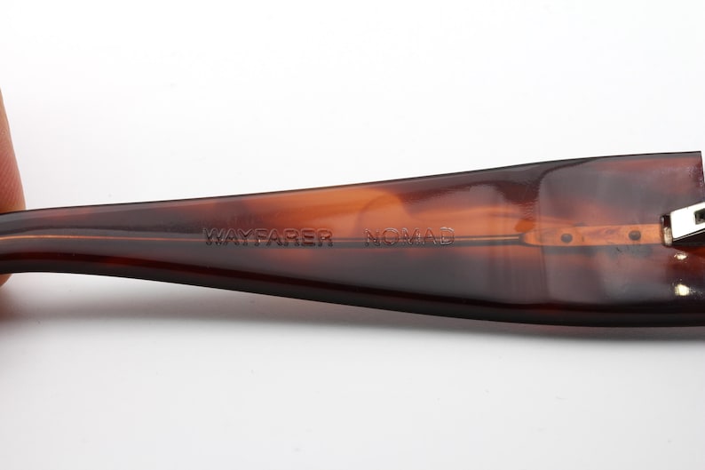 Puede incluir: Primer plano de una patilla de gafas Wayfarer Nomad marr&oacute;n. La patilla tiene un degradado de tonos marrones, con el texto "WAYFARER NOMAD" grabado en la montura. La patilla est&aacute; sujeta por los dedos de una persona.