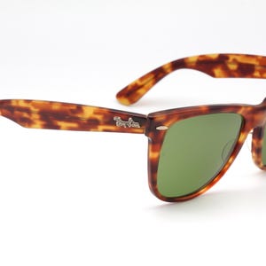May include: Tortoiseshell-patterned sunglasses with green lenses. The frames have a classic design with a subtle logo on the temple. The sunglasses are set against a white background.