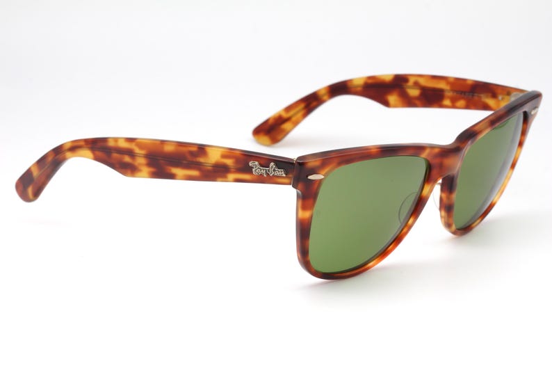 May include: Tortoiseshell-patterned sunglasses with green lenses. The frames have a classic design with a subtle logo on the temple. The sunglasses are set against a white background.