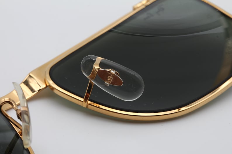 Op de afbeelding: Close-up van een goudkleurige zonnebril met donkere glazen. De zonnebril heeft een doorzichtig neuskussentje met een gouden accent en een klein embleem. Het montuur is glanzend goudkleurig.