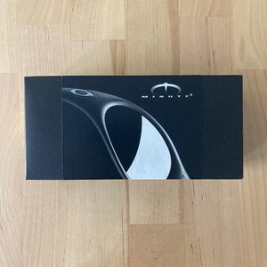 May include: A black rectangular box with the word "MINUTE" printed in white. The box features a curved design and a white logo. The box is on a wooden surface.