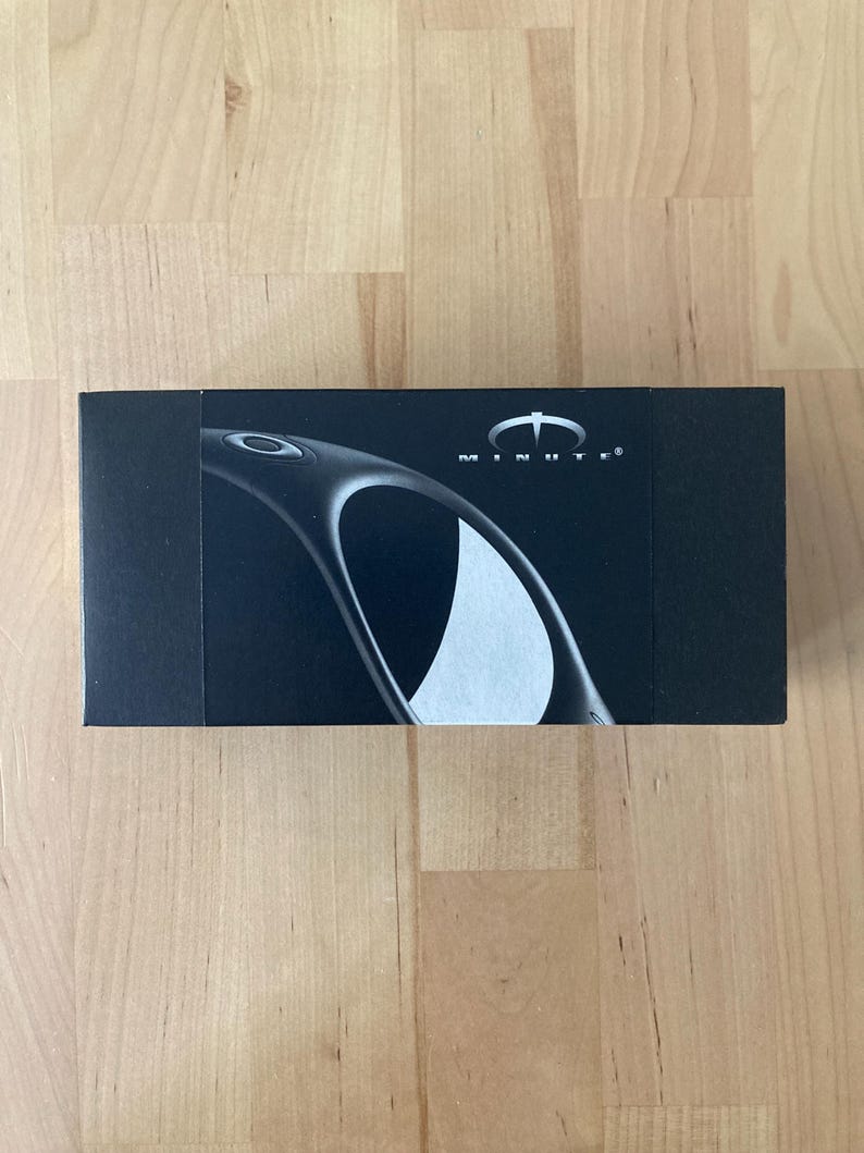 May include: A black rectangular box with the word "MINUTE" printed in white. The box features a curved design and a white logo. The box is on a wooden surface.