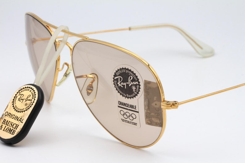 May include: Gold-tone Ray-Ban aviator sunglasses with brown lenses. The sunglasses have a white cord attached to the left arm. The sunglasses have a sticker on the right lens that says "100% UV protection Sunglasses by Bausch & Lomb Ray-Ban CHANGEABLE". The sunglasses also have a black and gold sticker on the left arm that says "ORIGINAL BAUSCH & LOMB".