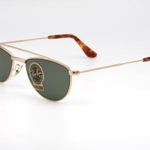 May include: Gold-framed sunglasses with green lenses. The sunglasses feature a double bridge and brown tortoiseshell-patterned temples. The Ray-Ban logo is visible on the lens. A classic eyewear accessory.