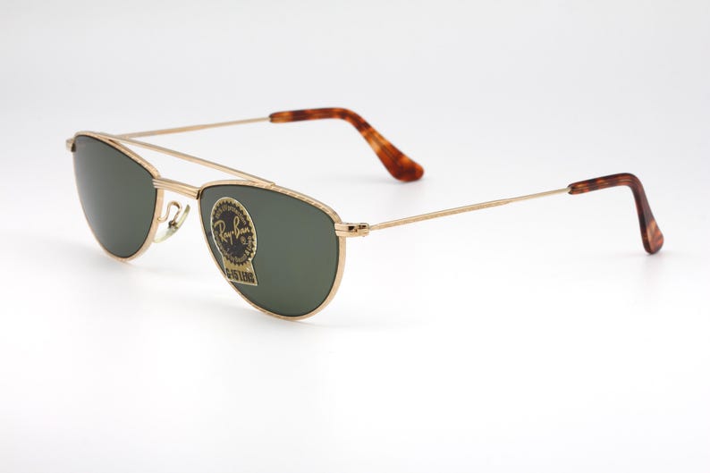 May include: Gold-framed sunglasses with green lenses. The sunglasses feature a double bridge and brown tortoiseshell-patterned temples. The Ray-Ban logo is visible on the lens. A classic eyewear accessory.