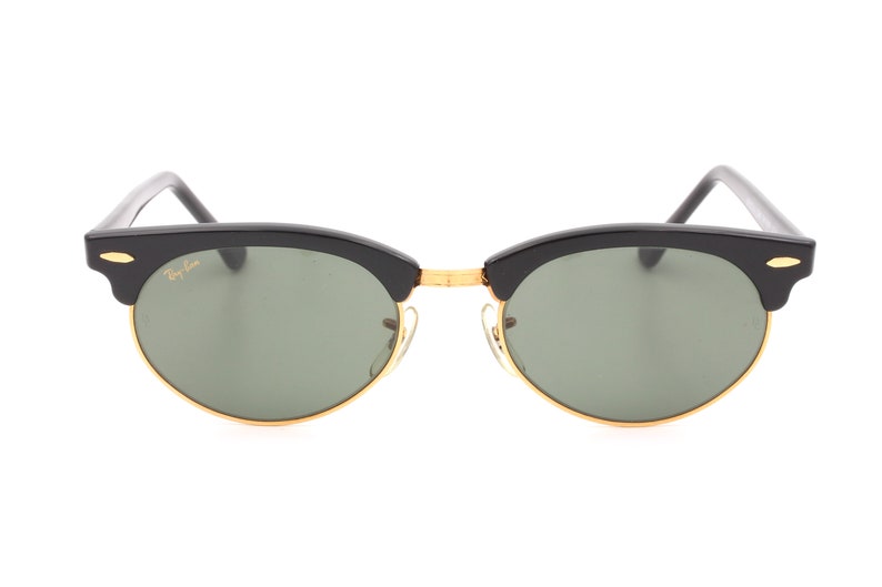 May include: Black Ray-Ban sunglasses with green lenses and gold accents. The sunglasses have a classic aviator style with a rounded shape.