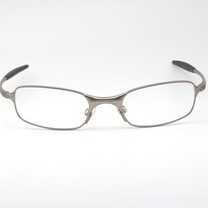 May include: Silver-toned eyeglasses with rectangular lenses and a thin metal frame. The temples have a curved design with black tips. The glasses are set against a plain white background.