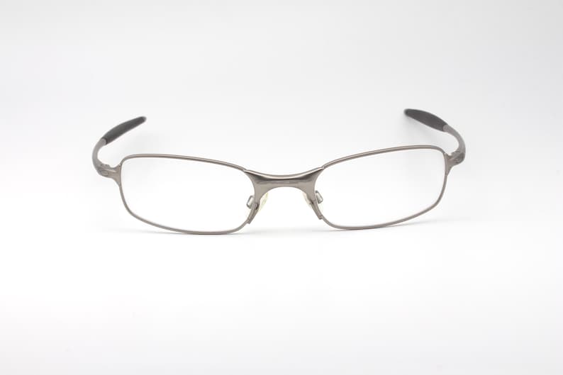 May include: Silver-toned eyeglasses with rectangular lenses and a thin metal frame. The temples have a curved design with black tips. The glasses are set against a plain white background.