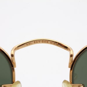 May include: Close-up of the gold-tone metal frame of a pair of Ray-Ban sunglasses. The frame is inscribed with "B&L RAY-BAN U.S.A."