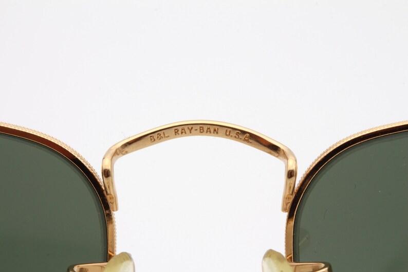 May include: Close-up of the gold-tone metal frame of a pair of Ray-Ban sunglasses. The frame is inscribed with "B&L RAY-BAN U.S.A."