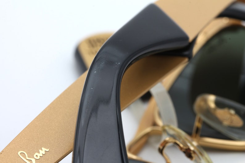Puede incluir: Primer plano de unas gafas de sol con montura dorada, patillas negras y lentes verdes. Las gafas tienen el logotipo de Ray-Ban en el lateral.