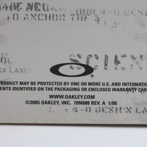 May include: A close-up of a white label with black text. The text reads "This product may be protected by one or more U.S. and international patents identified on the packaging or enclosed warranty card." The text also includes the website address "www.oakley.com" and the copyright information "&copy;2005 Oakley, Inc. 70N888 Rev. A 1/06".
