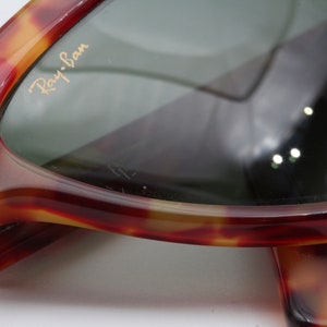 May include: Close-up of a pair of tortoiseshell Ray-Ban sunglasses. The sunglasses have a brown and gold tortoiseshell frame and green lenses. The Ray-Ban logo is visible on the right lens.