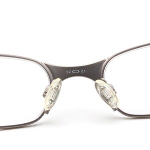 May include: Close-up of eyeglasses with a silver-colored frame. The bridge of the glasses has the numbers "52 21" printed on it. The nose pads are clear and rectangular.