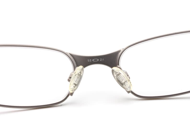 May include: Close-up of eyeglasses with a silver-colored frame. The bridge of the glasses has the numbers "52 21" printed on it. The nose pads are clear and rectangular.