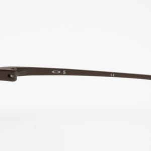 Op de afbeelding: Close-up van een bruin metalen brilmontuur. Het montuur heeft het nummer "5" en de merknaam "Oakley" op de poot gedrukt. De poot heeft ook de letters "CE" erop gedrukt.