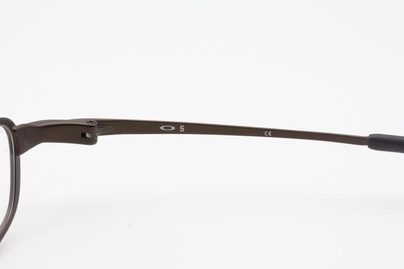 Op de afbeelding: Close-up van een bruin metalen brilmontuur. Het montuur heeft het nummer "5" en de merknaam "Oakley" op de poot gedrukt. De poot heeft ook de letters "CE" erop gedrukt.