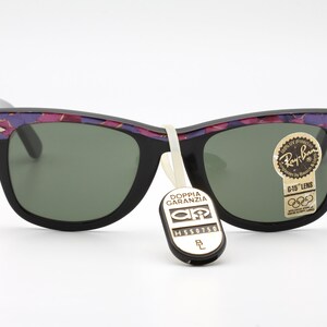 May include: Black Ray-Ban sunglasses with a purple and pink floral pattern on the frame. The sunglasses have green lenses and a G-15 lens tag. The sunglasses are also tagged with a "Doppia Garanzia" tag.