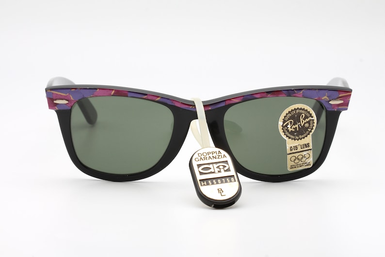 May include: Black Ray-Ban sunglasses with a purple and pink floral pattern on the frame. The sunglasses have green lenses and a G-15 lens tag. The sunglasses are also tagged with a "Doppia Garanzia" tag.