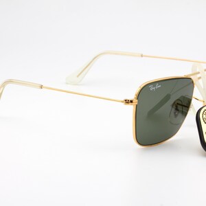 May include: Gold-framed square sunglasses with green lenses. The sunglasses are on a white surface with a black tag that reads "Original Bausch & Lomb".