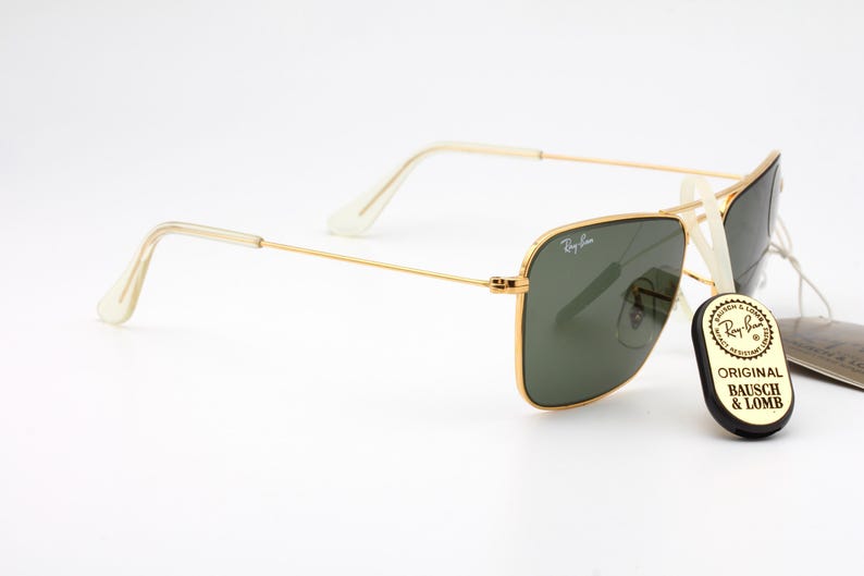 May include: Gold-framed square sunglasses with green lenses. The sunglasses are on a white surface with a black tag that reads "Original Bausch & Lomb".